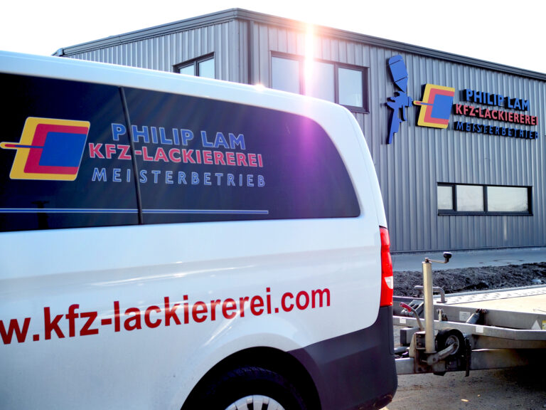 Gebäude der Kfz-Lackiererei mit Logo an der Hausfront, im Hintergrund. Im Vordergrund im Anschnitt Wagen der Lackiererei mit Logo auf dem Fenster, unten die Website. Anhänger angeschnitten.