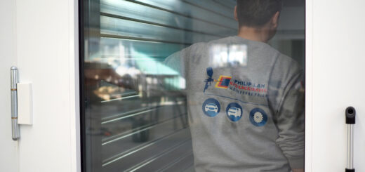 Mann im Vordergrund, durch die Fensterscheibe des Arbeitsraumes, von hinten, stehend sichtbar mit Sweat Shirt mit Logo der Lackiererei.